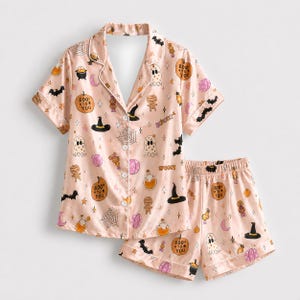 May include: A light pink pajama set consisting of a short-sleeved button-down shirt and matching shorts. The fabric features a repeating Halloween-themed print including ghosts, pumpkins with the text Boo to You, bats, spiderwebs, and hats.