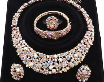 May include: A jewellery set featuring a wide, curved necklace, a hinged bracelet, a ring, and two stud earrings. Each piece is crafted from a gold-toned metal base and is densely encrusted with iridescent, multi-faceted crystals and rhinestones.