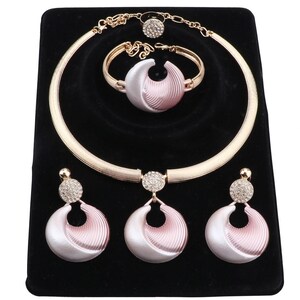 Op de afbeelding: Een sieradenset bestaande uit een goudkleurige choker-ketting, een armband, een ring en twee oorbellen. Elk stuk bevat een halvemaanvormige hanger met een roze en witte kleurverloop, geaccentueerd door kleine, heldere, ronde strass-steentjes.
