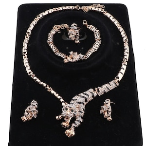 May include: A jewellery set featuring a rose gold-tone necklace, bracelet, ring, and stud earrings. Each piece is decorated with black enamel tiger stripes and clear rhinestones. The necklace features a large tiger pendant as the centrepiece.