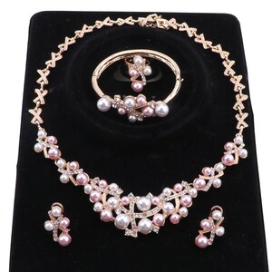 Puede incluir: Un conjunto de joyería en oro rosa que incluye collar, pulsera, anillo y pendientes. Cada pieza está adornada con perlas blancas y rosas y cristales transparentes, con una cadena de collar diseñada con un patrón de eslabones en forma de X.