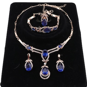 Può includere: Un set di gioielli composto da una collana, un bracciale e un paio di orecchini pendenti in tonalità oro rosa. Ogni pezzo è decorato con pietre blu intenso a forma di goccia e piccoli cristalli trasparenti con chiusura a moschettone.