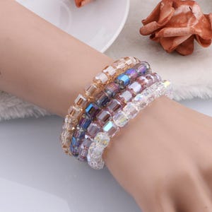 May include: Five beaded bracelets in a range of colours, including clear, gold, purple, and pink. The bracelets are made of square beads and are stacked on a wrist. The beads reflect light, creating a shimmering effect.