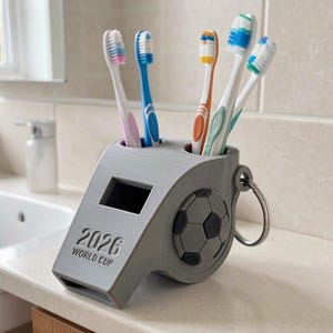 May include: A gray whistle-shaped toothbrush holder featuring a soccer ball design on the side and the text 2026 WORLD CUP embossed on the front. The holder contains four toothbrushes with white, blue, orange, and green bristles and handles.