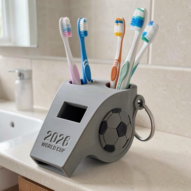 May include: A gray whistle-shaped toothbrush holder featuring a soccer ball design on the side and the text 2026 WORLD CUP embossed on the front. The holder contains four toothbrushes with white, blue, orange, and green bristles and handles.