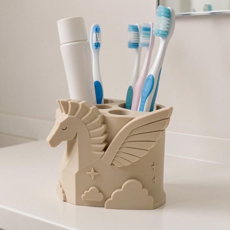 Op de afbeelding: Een beige cilindervormige tandenborstelhouder met een sculptuur van een pegasus met vleugels, wolken en stermotieven. De houder bevat een witte tube tandpasta en vier tandenborstels met blauwe, witte en roze borstelharen.