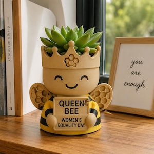 May include: A beige and yellow bee-shaped planter with a crown top, holding a sign that reads QUEEN BEE WOMEN'S EQUALITY DAY. The planter features black stripes, honeycomb-patterned wings, and a green succulent plant growing from the top opening.