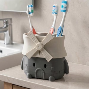 May include: A gray and cream-colored toothbrush holder shaped like a windmill with a textured brick-like base and a rotating blade design on the front. The holder features three circular openings at the top, currently containing three toothbrushes.