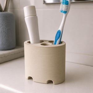 May include: A beige toothbrush holder with a ribbed design, holding a white tube of toothpaste and a white and blue toothbrush. The holder has four circular openings and sits on a white countertop. A gray container is visible in the background.