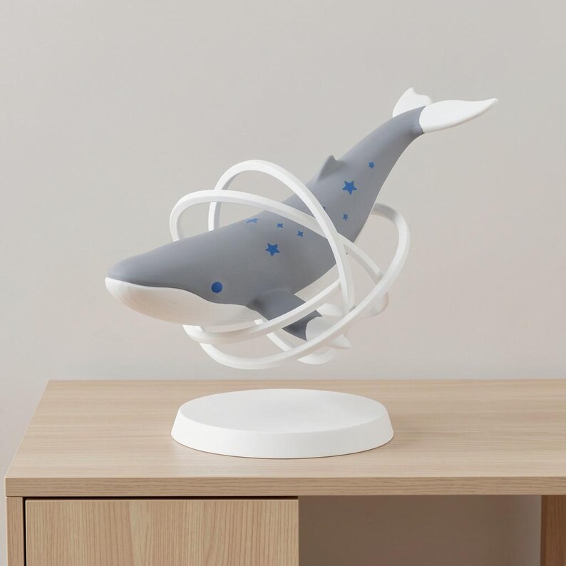 Op de afbeelding: Een grijze walvisfiguur met blauwe steraccenten en witte accenten, omcirkeld door witte ringen, lijkt boven een witte cirkelvormige basis te zweven. De walvis wordt getoond op een licht houten oppervlak.