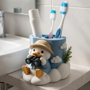 Puede incluir: Un soporte cil&iacute;ndrico para cepillos de dientes de color azul claro con un pato en 3D que lleva un sombrero beige y una chaqueta azul mientras sostiene unos binoculares negros. El soporte est&aacute; decorado con nubes blancas y contiene un tubo.