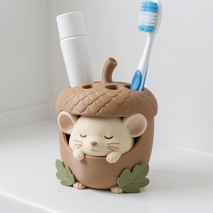 May include: A brown acorn-shaped toothbrush holder featuring a cream-colored mouse with closed eyes resting inside. The holder has a textured cap top with two circular openings, one containing a white tube and the other a blue and white toothbrush.