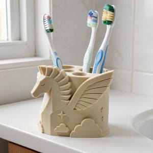 Op de afbeelding: Een beige tandenborstelhouder in de vorm van een gevleugeld paard, voorzien van gebeeldhouwde manen, vleugels en wolkenmotieven. De houder heeft drie ronde openingen aan de bovenzijde, met daarin drie tandenborstels met witte stelen en gekleurde haren.