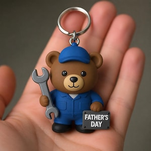 May include: A brown bear keychain wearing a blue cap and blue work uniform. The bear holds a grey wrench in one hand and a small grey rectangular sign in the other hand with the white text FATHER'S DAY printed on it. The keychain has a metal ring.