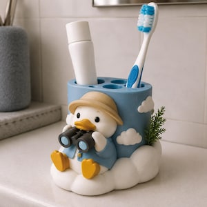 Puede incluir: Un soporte cil&iacute;ndrico azul para cepillos de dientes con un pato blanco que lleva un sombrero beige y una chaqueta azul mientras sostiene unos binoculares negros. La base tiene forma de nubes blancas y contiene un tubo de pasta dental y un cepillo.