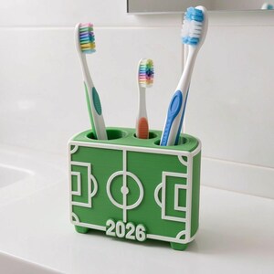 May include: A green toothbrush holder with a white soccer field design, holding three toothbrushes. The toothbrushes have white handles with green, blue, and orange accents, and colorful bristles. The number "2026" is printed on the front.