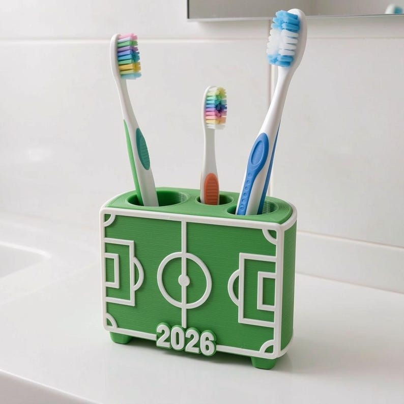 May include: A green toothbrush holder with a white soccer field design, holding three toothbrushes. The toothbrushes have white handles with green, blue, and orange accents, and colorful bristles. The number "2026" is printed on the front.