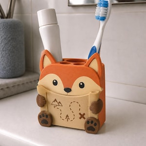 May include: A fox-shaped toothbrush holder in orange, cream, and brown, featuring a map graphic with mountain and tree icons. The holder has two circular openings on top, currently containing a white tube and a blue and white toothbrush.
