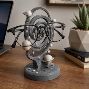 May include: A dark gray spiral-shaped glasses stand featuring white planetary spheres with rings and small star accents. The base is textured like a cratered moon surface. A pair of black-rimmed eyeglasses rests on the central spiral structure.