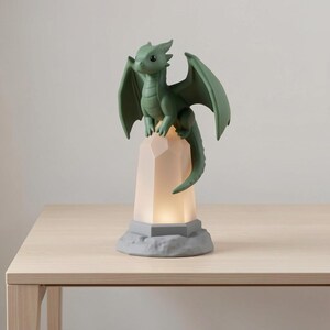 May include: A green dragon figurine with outstretched wings sits atop a light-up, faceted crystal-shaped base. The dragon is a muted green color, and the base emits a soft, warm glow. The base sits on a gray, textured platform. The figurine is on a wooden table.
