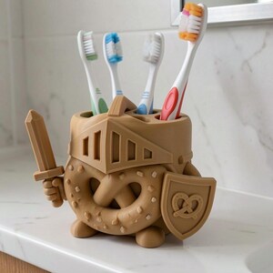 May include: A tan, knight-shaped toothbrush holder featuring a pretzel-textured body, a helmet visor, a sword in one hand, and a shield with a pretzel emblem in the other. The holder contains four toothbrushes with white, blue, green, and orange bristles.