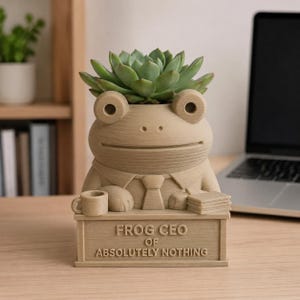 K&ouml;nnte beinhalten: Ein beigefarbener, 3D-gedruckter &Uuml;bertopf in Form eines Frosches mit Krawatte, der an einem Schreibtisch mit einer Tasse und einem Stapel Papier sitzt. Auf dem Schreibtisch steht "FROG CEO OF ABSOLUTELY NOTHING". Eine gr&uuml;ne Sukkulente ist eingepflanzt.