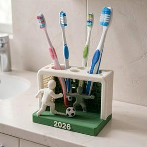 May include: A white and green toothbrush holder shaped like a football goal with a net, featuring a white player figure, a grey goalkeeper figure, a small football, and the year 2026 printed on the base, holding four colourful toothbrushes.