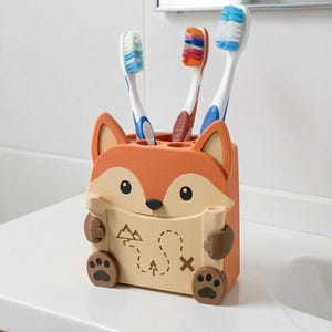 May include: A fox-shaped toothbrush holder in orange, cream, and brown, featuring a map graphic with mountains and a path. The holder contains three toothbrushes with blue, white, and multi-colored bristles, standing upright in circular slots.