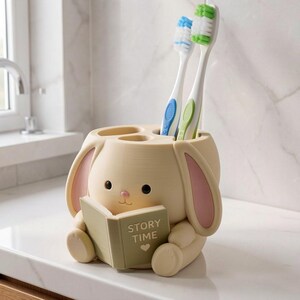 May include: A cream-colored rabbit-shaped toothbrush holder with pink inner ears, holding a small green book with the text STORY TIME and a heart icon. Two toothbrushes with blue and green bristles are inserted into the top of the holder.