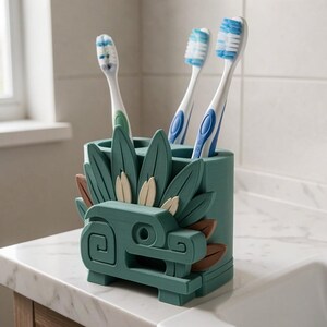 May include: A teal toothbrush holder featuring a stylized serpent design with layered leaf-like elements in green and tan. The holder has three circular openings at the top, currently containing three toothbrushes with blue and white bristles.