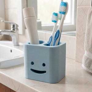 Puede incluir: Un porta cepillos de dientes azul claro con un dise&ntilde;o de cara sonriente. Contiene dos cepillos de dientes con cerdas azules y blancas, y un tubo de pasta de dientes. El soporte est&aacute; sobre una encimera blanca en un ba&ntilde;o.