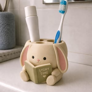 May include: A beige rabbit-shaped toothbrush holder featuring long ears with pink inner details, black eyes, and a small pink nose. The rabbit holds a green book with the text "STORY TIME" and a heart icon. It contains a white tube and a toothbrush.