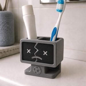 Puede incluir: Un soporte rectangular gris para cepillos de dientes con dos compartimentos, que presenta un panel frontal negro con un gr&aacute;fico blanco de una cara con el coraz&oacute;n roto, ojos en forma de X y un emblema de bal&oacute;n de f&uacute;tbol en la base.