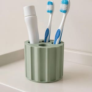May include: A sage green toothbrush holder with a ribbed design, holding two toothbrushes with blue accents and a tube of toothpaste. The toothbrushes have white bristles. The holder is on a white surface.