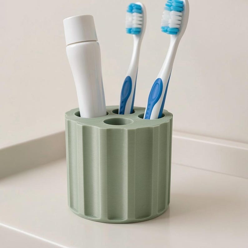 May include: A sage green toothbrush holder with a ribbed design, holding two toothbrushes with blue accents and a tube of toothpaste. The toothbrushes have white bristles. The holder is on a white surface.