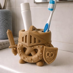 May include: A light brown, 3D-printed toothbrush holder shaped like a pretzel knight wearing a helmet. The figure holds a sword in one hand and a shield with a pretzel emblem in the other. It features two cylindrical openings for a toothbrush and toothpaste.