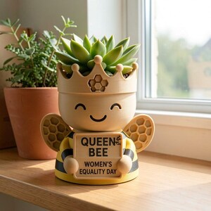 May include: A light yellow and black bee-shaped planter with a crown top holding a succulent. The planter features a sign that reads QUEEN BEE WOMEN'S EQUALITY DAY in black text. The design includes honeycomb patterns on the crown and wings.
