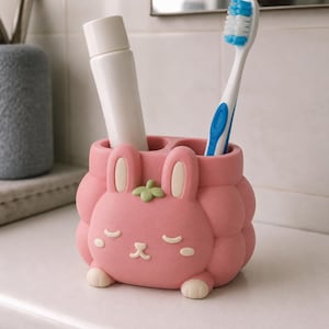 May include: A pink, rounded toothbrush holder shaped like a rabbit with closed eyes, a small green leaf on top, and two cream-colored paws at the base. The holder contains a white tube of toothpaste and a blue and white toothbrush with blue bristles.