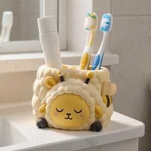 May include: A cream-colored toothbrush holder shaped like a sheep with a yellow face, black hooves, and antennae. It holds a white tube of toothpaste and two toothbrushes, one with yellow bristles and one with blue bristles, in its top slots.