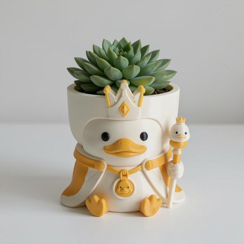 May include: A white and yellow planter shaped like a duck wearing a crown and cape. The duck holds a small scepter topped with a duck head and wears a medallion featuring a duck face. A green succulent plant is potted inside the duck-shaped container.