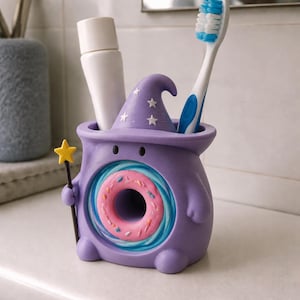 May include: A purple toothbrush holder shaped like a creature wearing a wizard hat with white stars. The center features a pink donut with colorful sprinkles surrounded by blue swirls. A small yellow star wand is attached to the side of the holder.