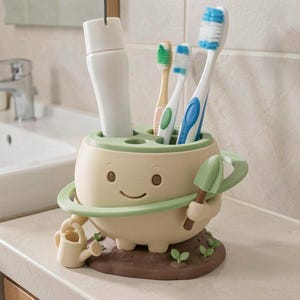 May include: A beige, round toothbrush holder shaped like a character with a smiling face, holding a small shovel and a watering can. The base is brown with small green sprouts. It contains a white tube and three toothbrushes with blue and green bristles.