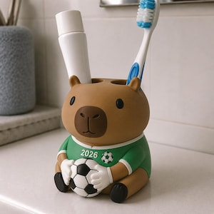 May include: A brown capybara-shaped toothbrush holder wearing a green soccer jersey with the text 2026 and a soccer ball graphic. The capybara is holding a black and white soccer ball in its hands. It contains a white tube and a blue toothbrush.