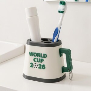 May include: A white and gray toothbrush holder with a green handle and the text "WORLD CUP 2026". The holder contains a tube of toothpaste and a blue and white toothbrush.