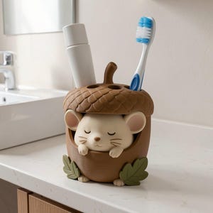 May include: A brown acorn-shaped toothbrush holder featuring a cream-colored mouse with closed eyes peeking out from the center. The holder has a textured cap, two green leaves at the base, and contains a white tube and a blue and white toothbrush.