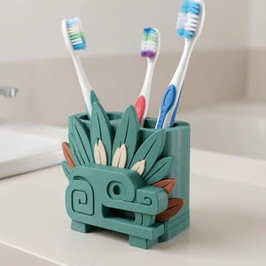 May include: A teal toothbrush holder featuring a stylized serpent design with layered leaf-like elements in teal, cream, and brown. The holder contains three toothbrushes with white handles and blue, green, and purple bristles.