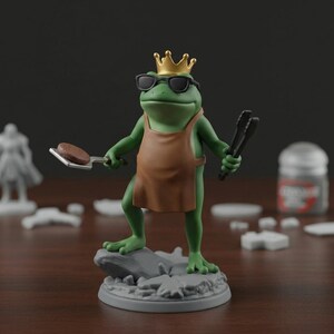 May include: A green frog figurine wearing sunglasses, a gold crown, and a brown apron, holding a burger on a spatula and tongs. The frog stands on a gray rock base, with a dark background and a wooden surface.