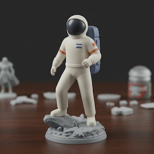 May include: A white astronaut figurine with a black helmet and blue backpack stands on a gray rocky base. The astronaut's suit has orange stripes on the arms and two blue stripes on the chest. Other miniature figures and a container are in the background.