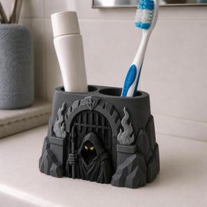 Puede incluir: Un soporte para cepillos de dientes gris oscuro con forma de puerta de fortaleza de piedra con una figura encapuchada que sostiene una lanza. La figura tiene ojos amarillos brillantes. La estructura tiene dos aberturas para un tubo y un cepillo.