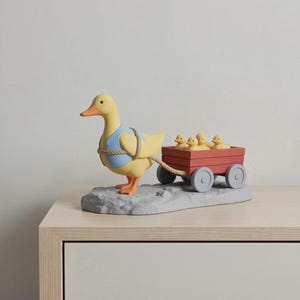 May include: A yellow duck figurine wearing a blue vest pulls a red wagon with five yellow ducklings. The duck is standing on a gray base. The wagon has gray wheels and is connected to the duck with a rope.